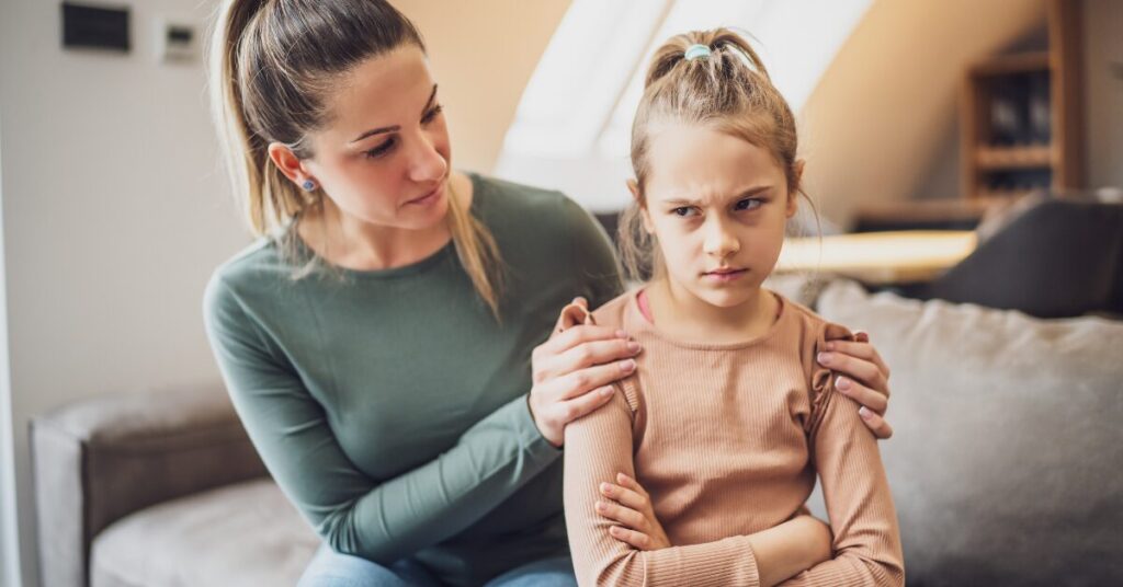 attune to child's perspective caucasian woman sits behind young girl with her hands on her shoulders in gentle touch. Girl has scowling face and arms crossed.