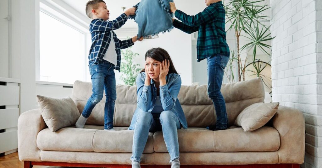 parent looking overwhelmed with head in hands as two kids have aragument over pillow behind her