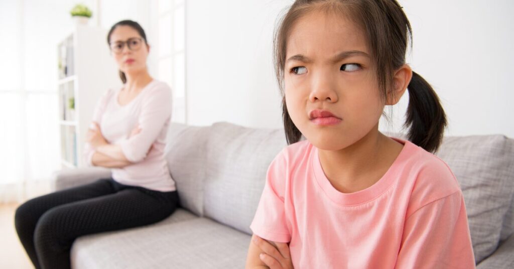 little girl scowling and mother in background with arms crossed staring at the back of her head