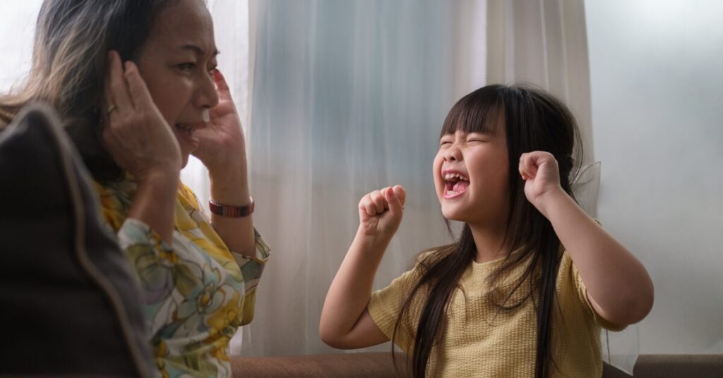 trauma reactions asian woman with hands over ears, exasperated expression on her face as a preschool age young asian girl screams with her eyes closed and hands in a fist depicting hyperrousal