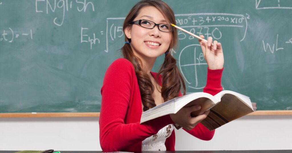 figure it out An Asian woma with glasses smiling holding a book and holding a pencil to her temple as she depicts she is navigating the uncertainty of this challenge