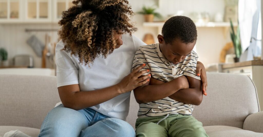 mother sitting on couch with boy both are looking down, mother has her arm around boy whose arms are crossed against his chest. he looks distressed