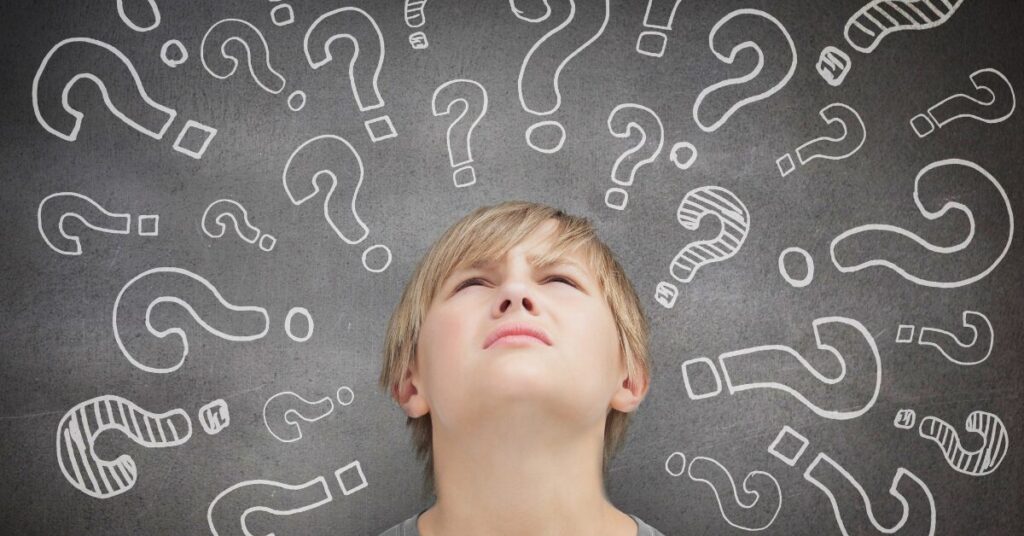 young boy in front of chalkboard with a bunch of question marks drawn on it. boy is looking up as if confused