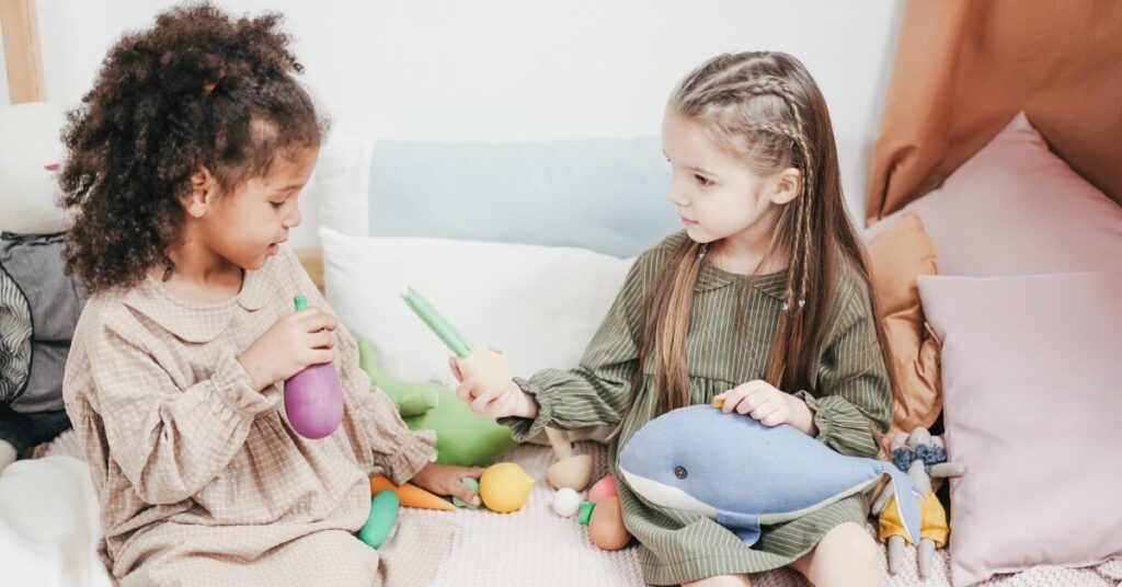 trauma reactions an african american young girl sitting on a couch with a caucasian preschool age girl offering her a toy. the african american girl appears to be hesitant to accept the toy. this is an example of both pleasing and appeasing and going along to get along