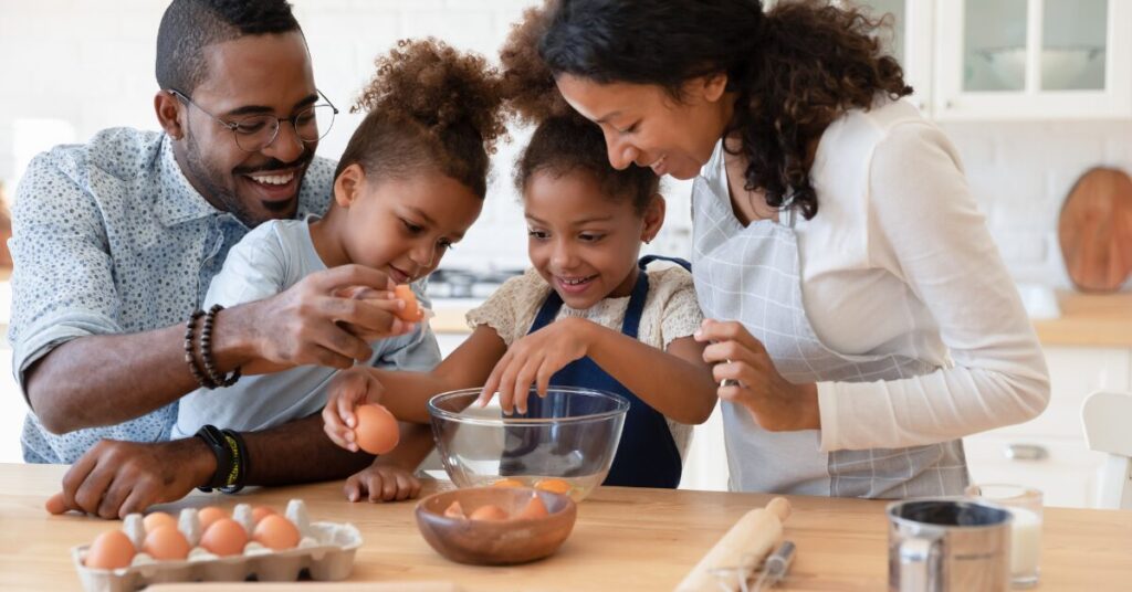 involving african american mom, dad, and two little girls involved in cooking something. the parents are involving the girls as the girls are cracking eggs into a bowl