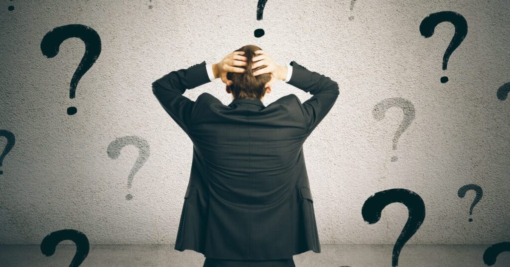 challenging behavior man in suit from behind with hands on back of head looking at background with a bunch of question marks to depict he is uncertain